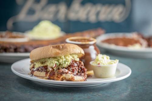 A close-up of a pulled pork sandwich topped with coleslaw, served with a side of coleslaw and barbecue sauce.