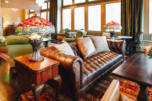 Cozy lounge with a brown leather sofa, decorative pillows, and stained glass lamps, featuring large windows and elegant decor.