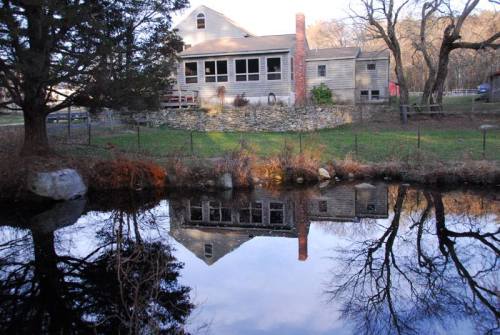 A house with a stone wall reflected in a calm pond, surrounded by trees and a grassy area.