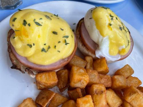 Two eggs Benedict on English muffins with ham, topped with hollandaise sauce, served with golden-brown diced potatoes.