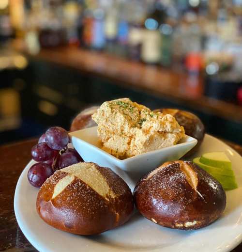 A plate with pretzel bites, a bowl of creamy dip, grapes, and apple slices, set against a blurred bar background.