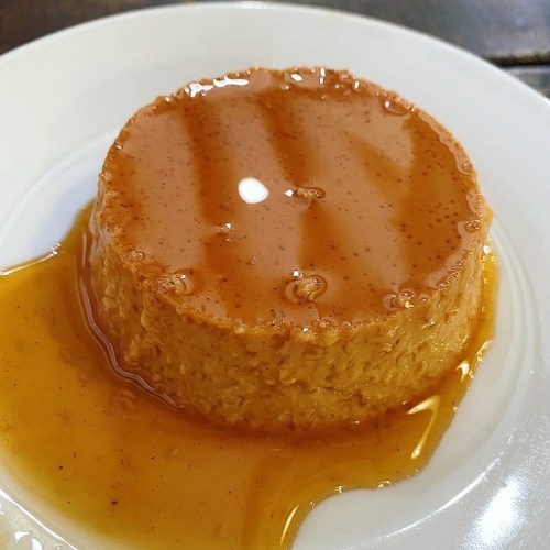 A round caramel flan on a white plate, topped with a glossy caramel sauce and surrounded by a light syrup.