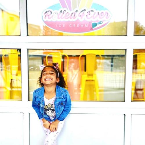 A smiling child in a denim jacket stands in front of a colorful ice cream shop window.