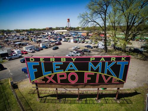 A large colorful sign for Derby Park Flea Market, overlooking a busy outdoor market with cars and vendors.