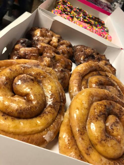 A box of assorted donuts, featuring glazed cinnamon rolls and sprinkled donuts in the background.