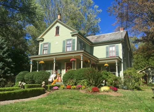 A charming green Victorian house surrounded by colorful flowers and lush greenery under a clear blue sky.
