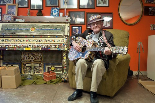 An elderly man in a colorful hat plays a guitar while sitting on a chair, surrounded by decorated walls and a unique piano.