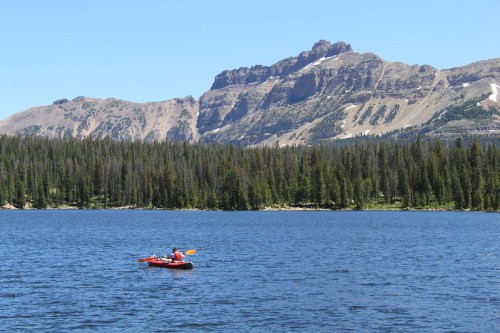 A person kayaking on a calm lake surrounded by mountains and dense forest under a clear blue sky.