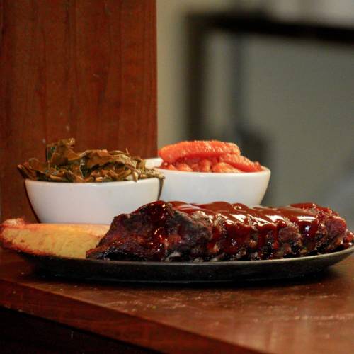 A plate of barbecue ribs with sauce, served with collard greens, pickled vegetables, and cornbread on the side.