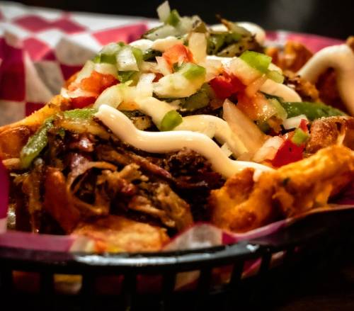 A close-up of a loaded waffle topped with pulled meat, diced vegetables, and drizzled with sauce in a basket.