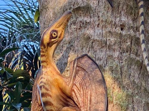A wooden sculpture of a dinosaur-like creature perched on a tree trunk, surrounded by greenery.