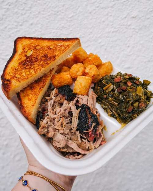 A hand holding a takeout tray with pulled pork, collard greens, tater tots, and two slices of toasted bread.