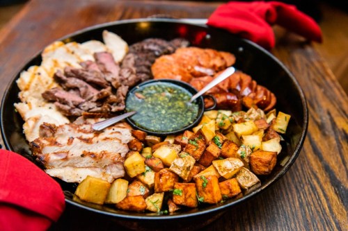 A large platter featuring assorted meats, roasted potatoes, and a small bowl of green sauce, garnished with herbs.