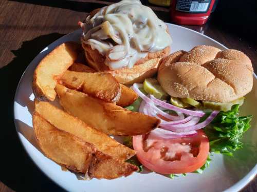 A plate with a cheesy sandwich, lettuce, tomato, pickles, onions, and crispy potato wedges on the side.