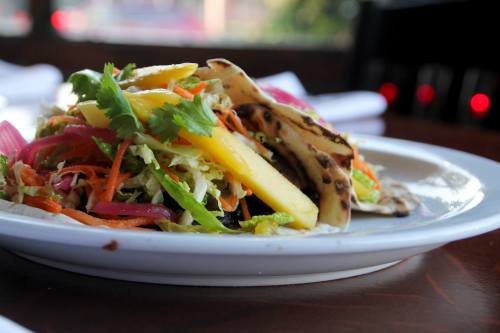 A plate of tacos topped with colorful vegetables and mango, garnished with cilantro, on a wooden table.