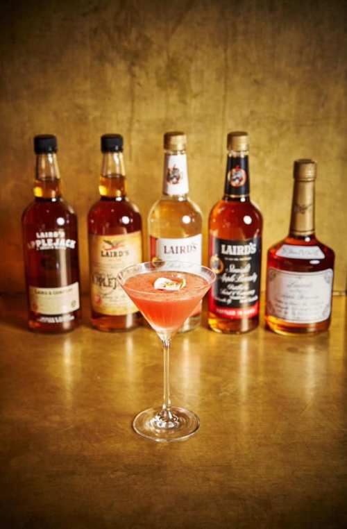 A pink cocktail in a martini glass, with several bottles of Laird's spirits in the background on a golden surface.
