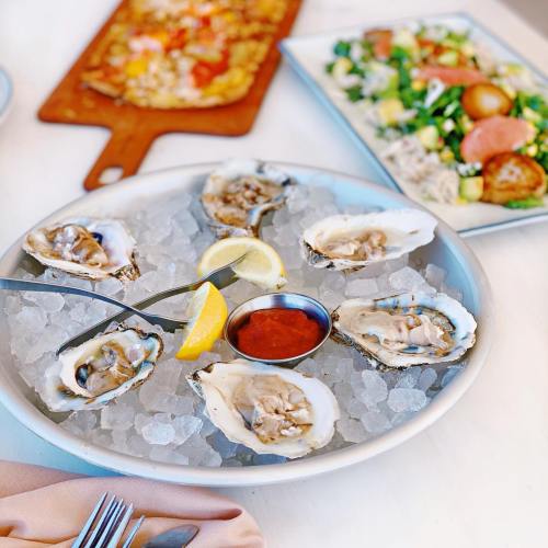 A platter of fresh oysters on ice with lemon wedges and cocktail sauce, surrounded by other dishes in the background.