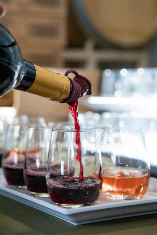A bottle of red wine is being poured into a glass, with other drinks visible on a tray in the background.