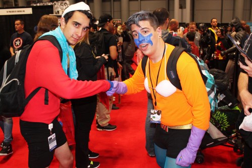 Two costumed individuals shake hands at a convention, surrounded by a crowd on a red carpet.