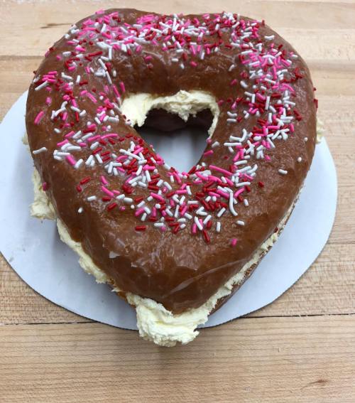 A heart-shaped donut with chocolate icing and pink and white sprinkles on a wooden surface.