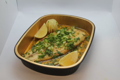 A baked fish fillet garnished with green onions and lemon slices in a gold-trimmed black tray.