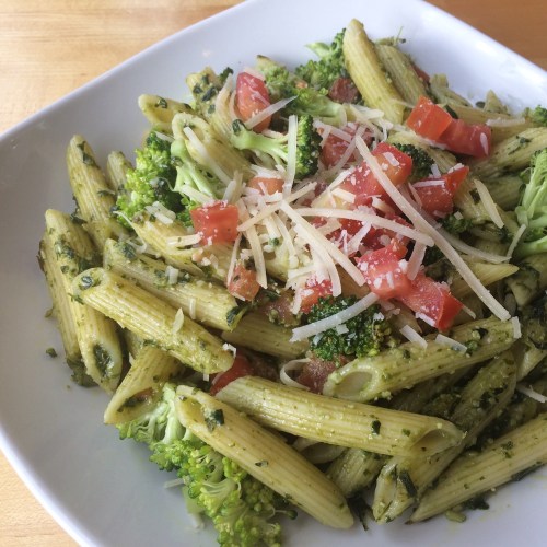 A bowl of penne pasta with pesto, broccoli, diced tomatoes, and grated cheese on top.