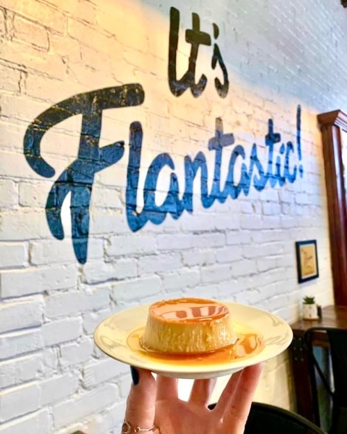 A hand holds a flan on a plate in front of a wall with the text "It's Flantastic!" painted in blue.