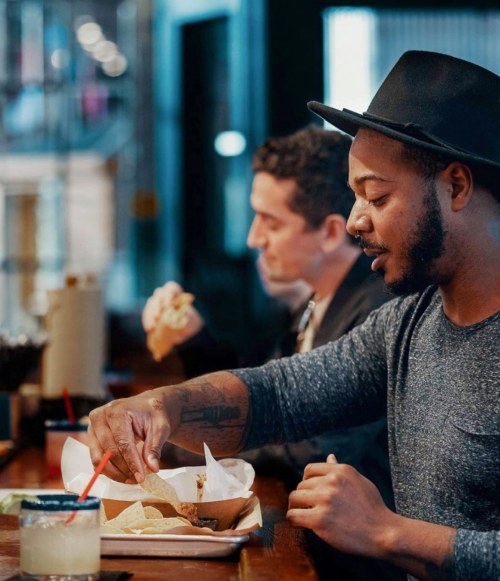 A man in a hat enjoys food at a bar, while another man eats in the background. Drinks are on the table.