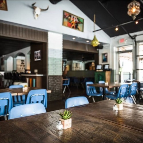 Interior of a restaurant featuring wooden tables, blue chairs, and decorative plants, with art on the walls.