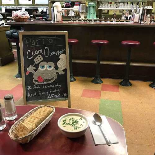 A bowl of clam chowder on a table with a basket of crackers, and a chalkboard sign advertising the special.