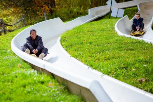 Two children enjoy a summer day on a winding slide, surrounded by green grass and autumn foliage.