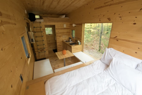 Cozy wooden interior of a tiny cabin with a bed, kitchenette, and large windows overlooking a forest.