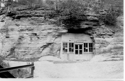 A weathered wooden building set against a rocky cliff, with a dirt path leading up to it.