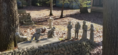 A whimsical outdoor scene featuring stone sculptures of people gathered around a small stage in a wooded area.