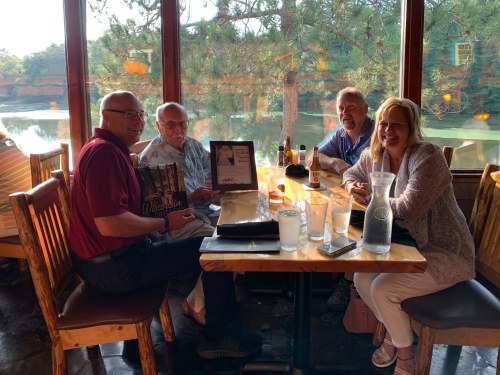 Four people sitting at a table in a restaurant, smiling and holding menus, with a scenic view of trees and water outside.