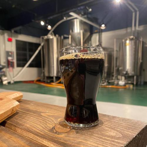 A glass of dark beer sits on a wooden table, with brewing equipment visible in the background.