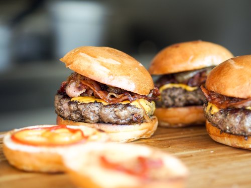 Three delicious burgers with bacon and cheese on toasted buns, with two open buns and condiments on a wooden board.