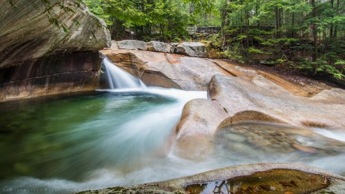 A serene river flows over smooth rocks, surrounded by lush green trees in a tranquil natural setting.