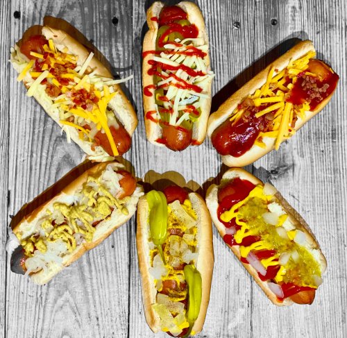 A variety of hot dogs arranged in a star pattern, topped with cheese, ketchup, mustard, and pickles on a wooden surface.