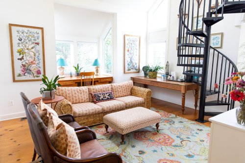 Cozy living room with a spiral staircase, floral artwork, a patterned sofa, and a wooden desk by large windows.