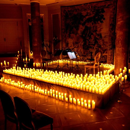 A dimly lit room filled with glowing candles, creating a warm ambiance around a small stage with chairs and sheet music.