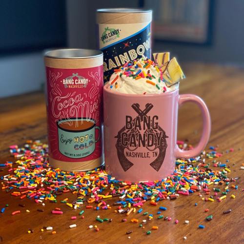 A pink mug with whipped cream and sprinkles, surrounded by colorful candy containers on a wooden table.