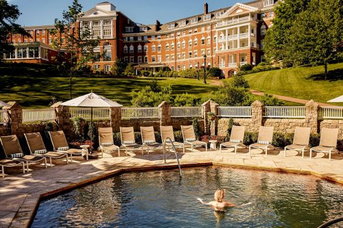 A serene pool area with lounge chairs, surrounded by greenery and a large historic building in the background.