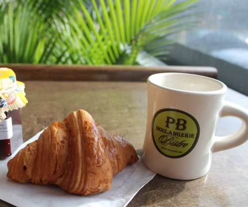 A croissant next to a coffee mug on a table, with greenery in the background.
