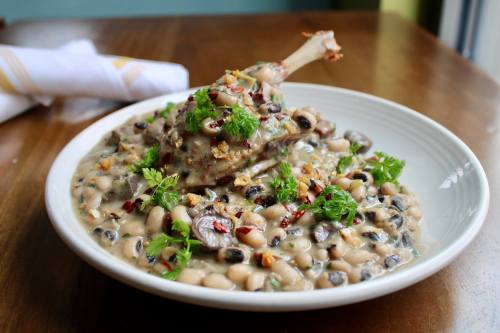 A bowl of creamy black-eyed peas topped with herbs, spices, and a piece of meat on a wooden table.