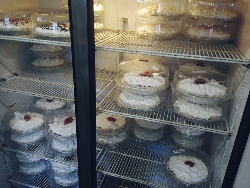 A refrigerator filled with multiple pies topped with whipped cream and cherries, neatly arranged in plastic containers.
