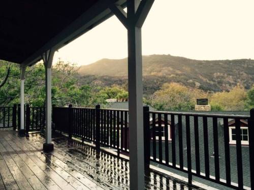 A wooden deck with a railing overlooks a misty mountain landscape at sunset, with wet planks reflecting light.