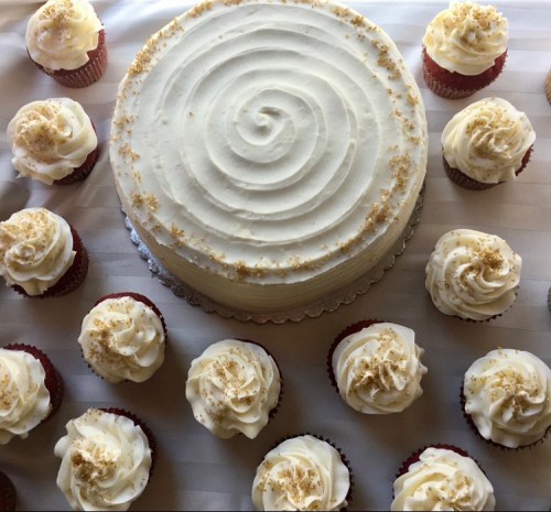 A round white cake with a spiral design surrounded by red velvet cupcakes topped with cream cheese frosting.