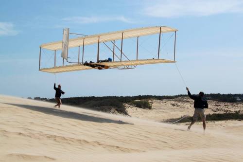 Two people on a sandy dune are launching a vintage-style glider into the air.