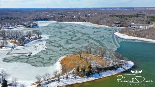 Aerial view of a frozen lake with swirling ice patterns, surrounded by trees and a snowy landscape.